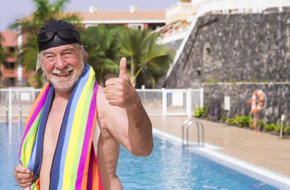 Smiling senior man with white hair comes out of the pool wearing swimming cap and goggles gesturing ok with hand - with a colorful towel on his shoulders - active seniors in retirement