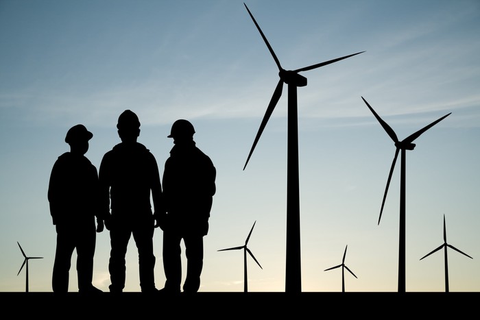 Three workers are shown in silhouette near six wind turbines.